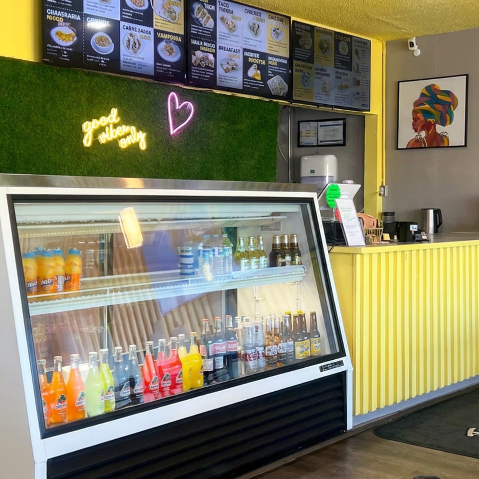Bright yellow restaurant interior with illuminated menu boards, colorful bottled drinks, beverage counter, and vibrant wall portraits.