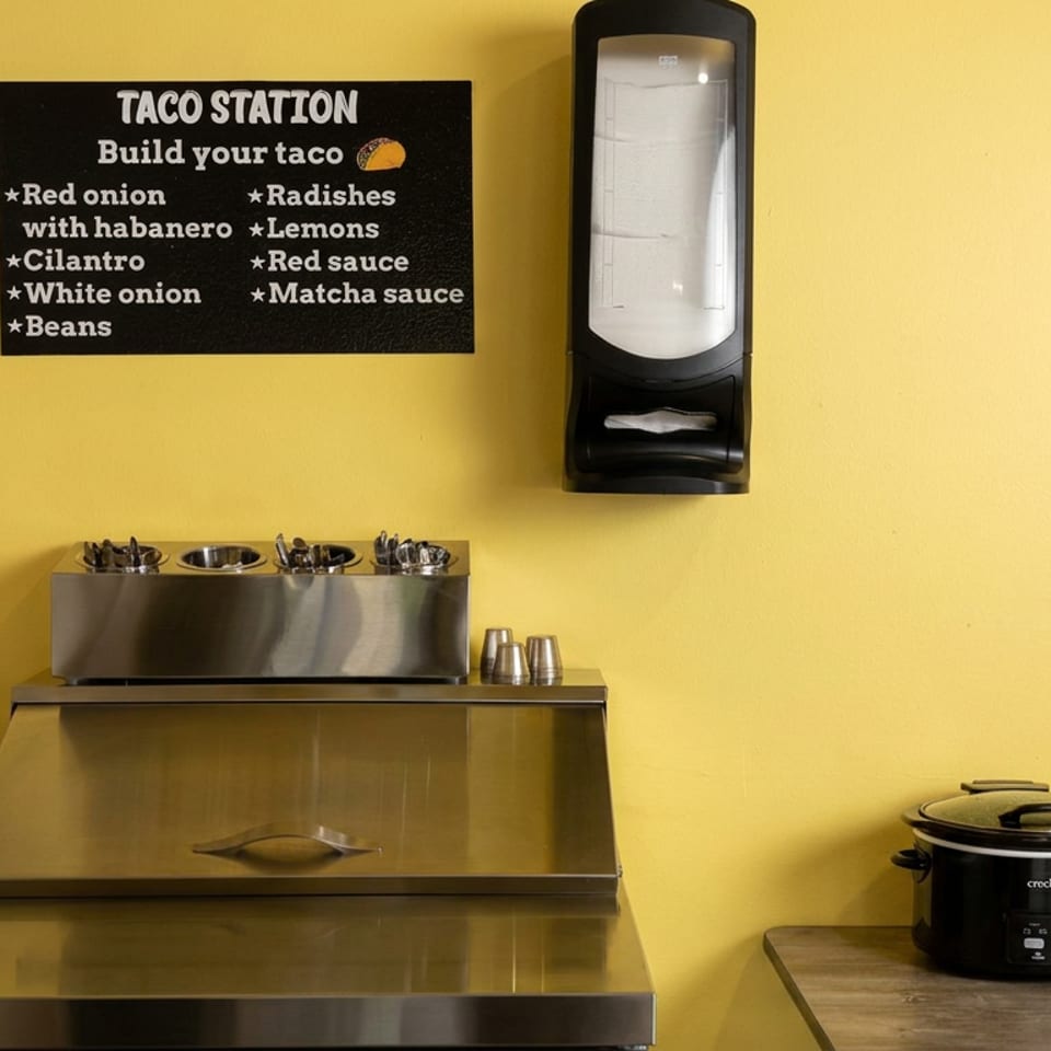 Taco station with build-your-own toppings sign, stainless serving trays, utensil holder, napkin dispenser, and crockpot against a yellow wall.